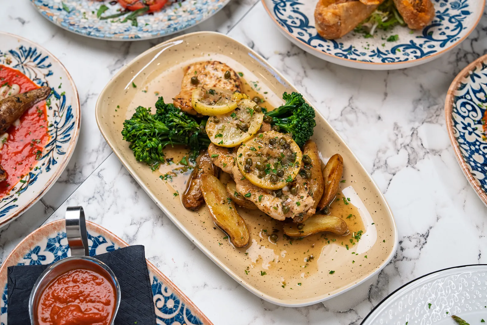 Chicken piccata with lemon slices, capers, fingerling potatoes and broccolini on a cream platter